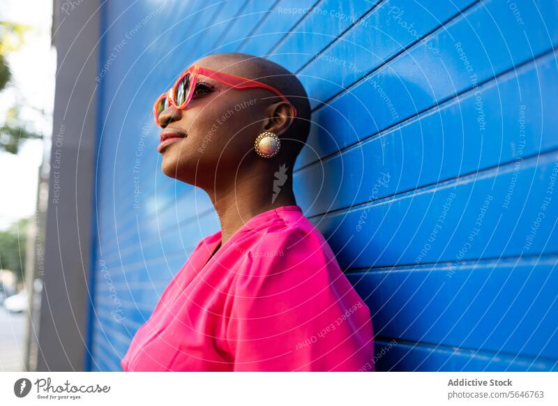 Happy Stylish schwarze Frau mit rasiertem Kopf in Sonnenbrille und rosa Kleid stehen gegen blaue Wand selbstbewusst cool trendy Outfit Individualität Stil