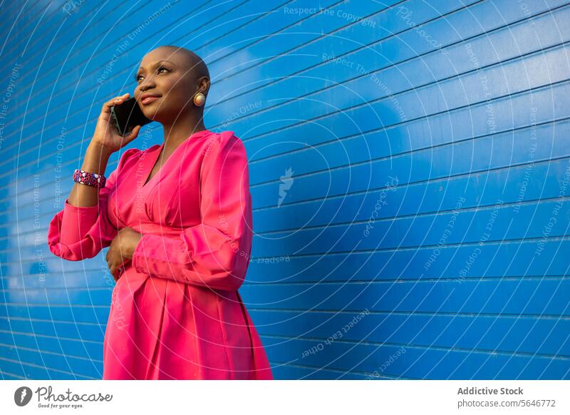 Stilvolle schwarze Frau mit rasiertem Kopf in rosa Kleid spricht mit Handy gegen blaue Wand Smartphone benutzend Telefonanruf reden Glück kahl Gespräch sprechen