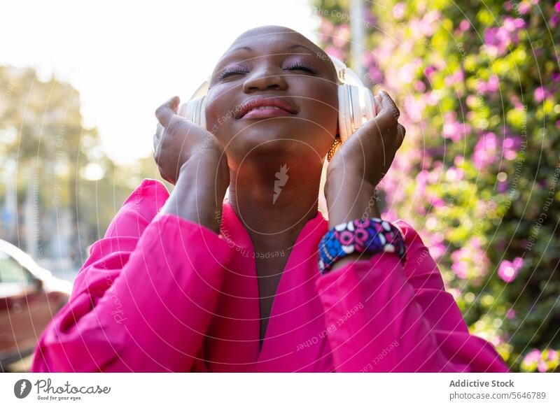 Glückliche schwarze Frau mit rasiertem Kopf im rosa Kleid hört Musik über Kopfhörer mit geschlossenen Augen im Park zuhören Drahtlos benutzend sorgenfrei trendy