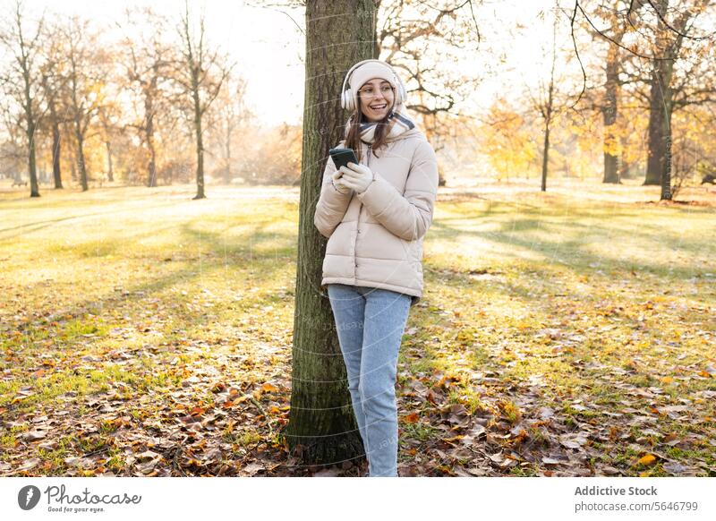 Fröhliche junge Frau in warmer Kleidung mit Smartphone im Park zuhören Musik Kopfhörer benutzend Herbst warme Kleidung positiv Lächeln Mobile Glück Gerät