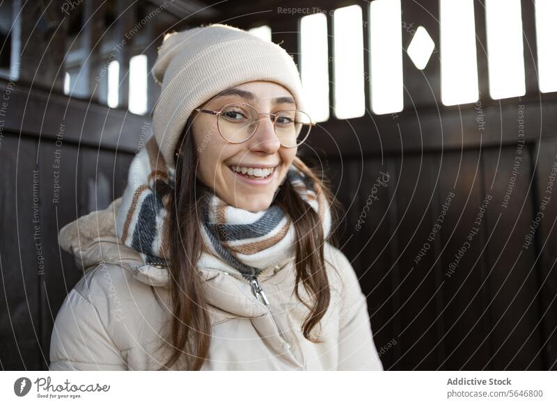 Lächelnde junge Frau in warmer Jacke an einem Holzzaun stehend Winter Herbst warme Kleidung positiv Glück Hut Porträt Brille froh Verschlussdeckel Freude Surfen