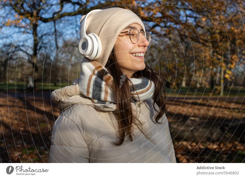 Lächelnde Frau mit Kopfhörern, die im Herbst im Park Musik hört zuhören Gesang positiv genießen Freizeit warme Kleidung Brille Drahtlos jung Glück Hut