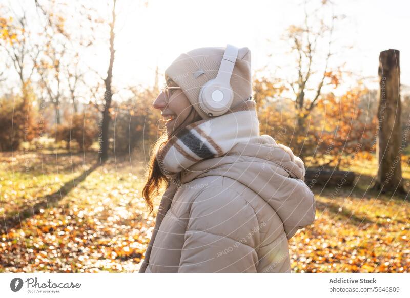 Lächelnde Frau mit Kopfhörern, die im Herbst im Park Musik hört zuhören Gesang positiv genießen Freizeit warme Kleidung Brille Drahtlos jung Glück Hut