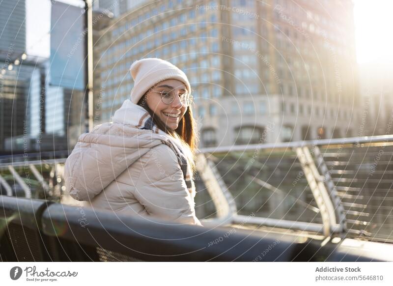 Lächelnde Frau in warmer Kleidung sitzt auf einer Metallbank am Geländer in der Stadt Straße metallisch Bank Großstadt Gebäude modern Reling warme Kleidung
