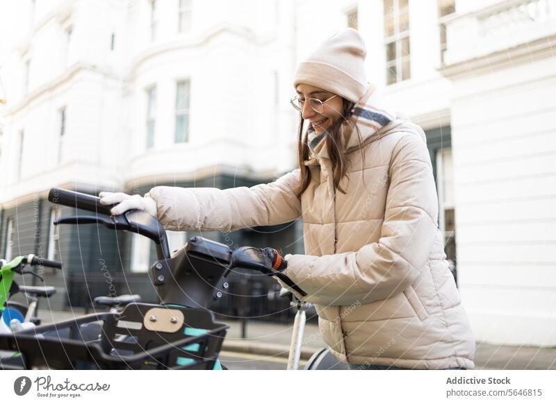Lächelnde Frau in gemütlicher Jacke mit Elektrofahrrad auf der Straße elektrisch Tretroller Großstadt modern Gebäude trendy positiv warme Kleidung Stil Fassade