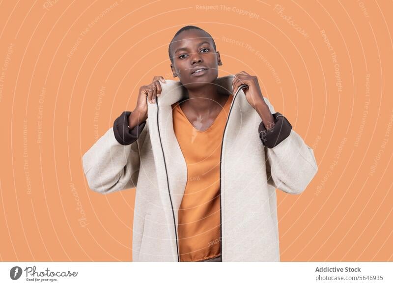 Porträt einer jungen, mutigen Afroamerikanerin in einem langen Mantel mit Reißverschluss, die selbstbewusst in die Kamera blickt und vor einem orangefarbenen Hintergrund steht