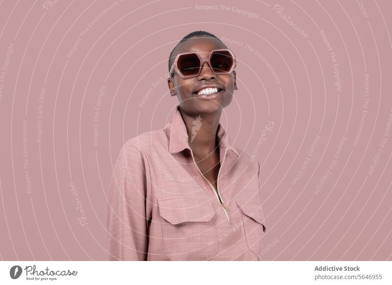 Junges, fröhliches, kühnes afroamerikanisches weibliches Modell in stilvoller rosa Kleidung und Sonnenbrille, das vor einem rosa Hintergrund steht und wegschaut