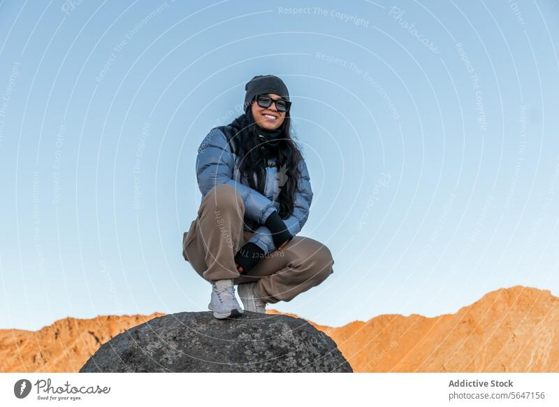 Glückliche Wanderin, die in warmer Kleidung und mit Sonnenbrille bei Tageslicht auf einem Felsen sitzend in die Kamera schaut Frau Reisender Tourist Lächeln