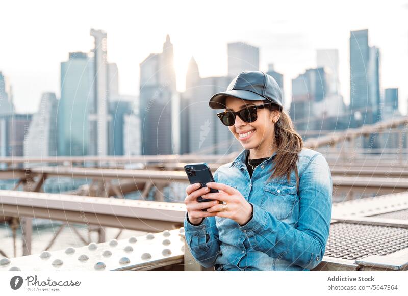 Frau mit Smartphone vor Manhattan-Kulisse Sonnenbrille Verschlussdeckel Lächeln Skyline Stadtbild New York State im Freien urban Brücke Mode Freizeit lässig
