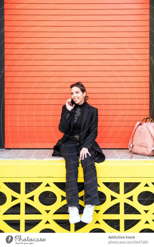 Junge Frau sitzt an einer orangefarbenen Wand und spricht mit ihrem Smartphone reden Telefonanruf sprechen Lächeln Glück Straße Gespräch heiter jung Mobile