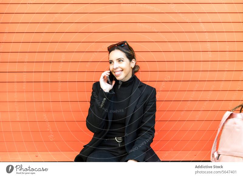 Junge Frau sitzt an einer orangefarbenen Wand und spricht mit ihrem Smartphone reden Telefonanruf sprechen Lächeln Glück Straße Gespräch heiter jung Mobile