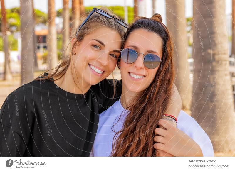 Lächelnde kaukasische Freundinnen mit Sonnenbrille Frauen Porträt Glück Park Zusammensein modisch Bonden Kopfschuss Unschärfe Arme ringsherum brünett blond