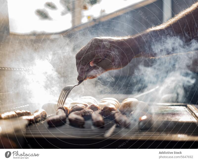 Unbekannter Mann grillt Würstchen auf einem mit Rauch bedeckten Grillrost Küchenchef Koch stoßen Gabel vorbereiten Ablage Wurstwaren Barbecue männlich
