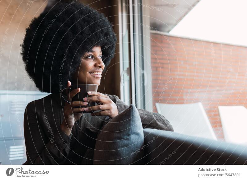 Fröhliche schwarze Frau mit einer Tasse Kaffee Heißgetränk Getränk Afro-Look Fenster Lächeln Freizeit Erfrischung Frisur feminin Vorschein Glück charmant