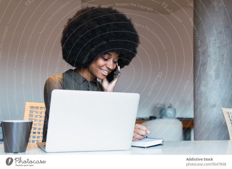 Glückliche schwarze Frau schreibt in ein Notizbuch neben einem Laptop Arbeit freiberuflich Arbeitsplatz Telefonanruf Notebook zur Kenntnis nehmen sprechen