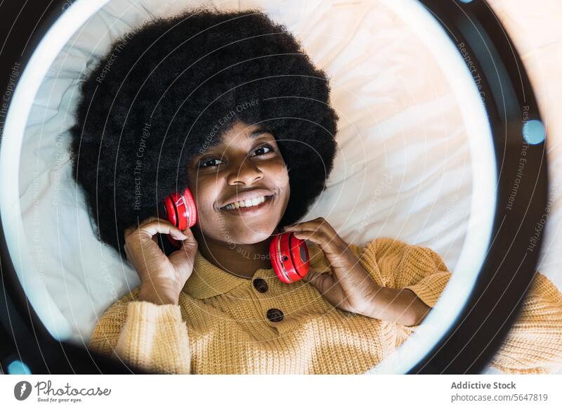 Lächelnde schwarze Frau, die mit Kopfhörern Musik hört zuhören benutzend Bett ruhen heimwärts Wochenende Kälte Afroamerikaner Afro-Look Apparatur Ringlampe