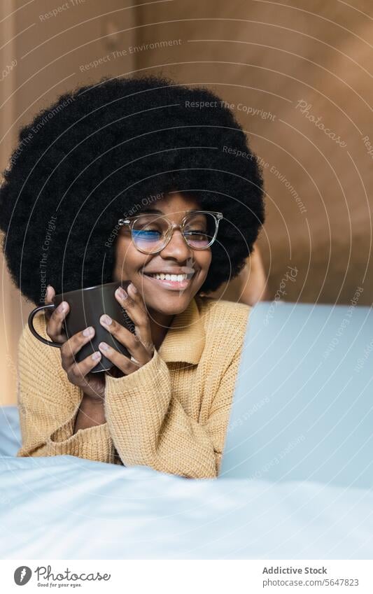 Lächelnde schwarze Frau mit Laptop und Kaffee im Bett benutzend ruhen sich[Akk] entspannen Wochenende Freizeit heimwärts Afroamerikaner Afro-Look zuschauen