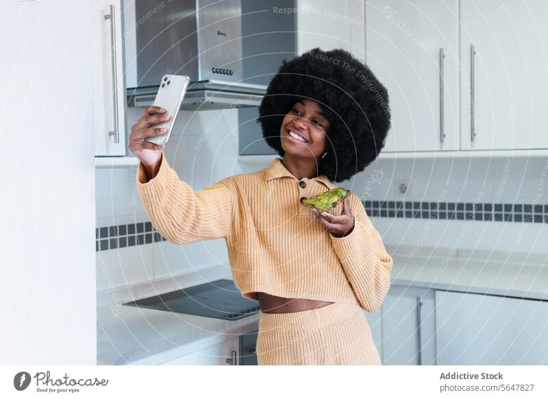Lächelnde schwarze Frau macht Selfie mit Toast Frühstück Gesunder Lebensstil Küche Smartphone Selbstportrait Avocado benutzend Morgen Afroamerikaner Afro-Look