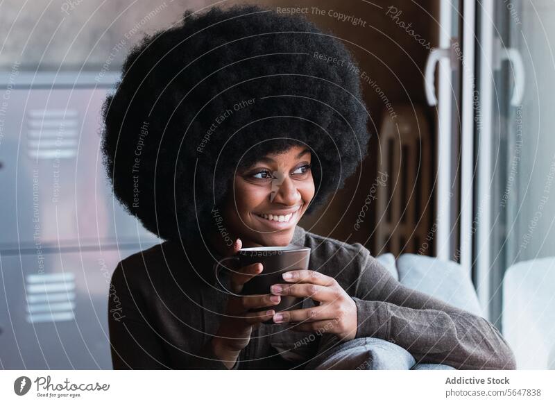 Fröhliche schwarze Frau mit einer Tasse Kaffee Heißgetränk Getränk Afro-Look Fenster Lächeln Freizeit Erfrischung Frisur feminin Vorschein Glück charmant