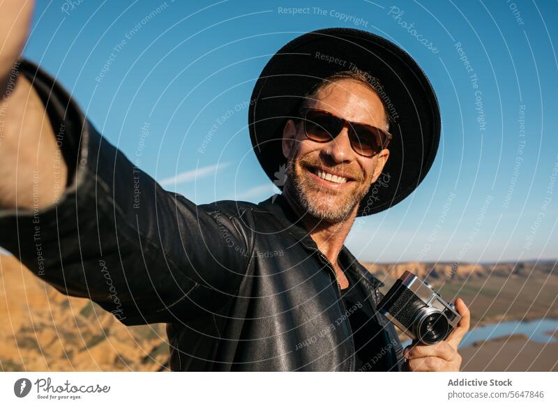 Lächelnder Reisender nimmt Selfie mit Kamera auf Abenteuer Abenteurer Fotograf Fotokamera reisen Erkundung wüst malerisch Glück heiter Mann Hut Sonnenbrille