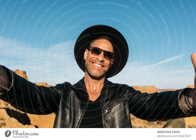 Fröhlicher Mann, der ein Selfie mit ausgestreckten Armen macht und die Natur genießt Lächeln Glück Freude im Freien Hut Sonnenbrille schwarze Jacke Leder