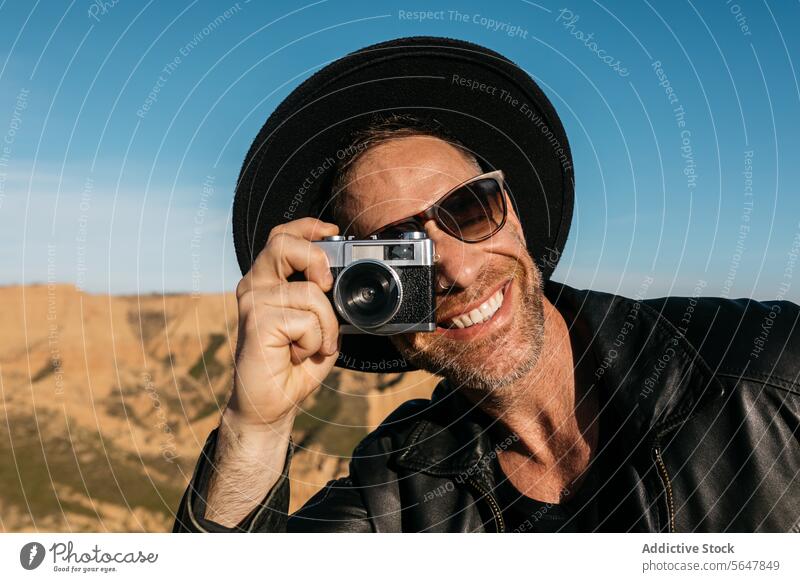 Lächelnder Fotograf, der den Moment im Freien mit einer Filmkamera festhält Mann Fotokamera altehrwürdig Hut malerisch Hintergrund freudig unter Leder Jacke