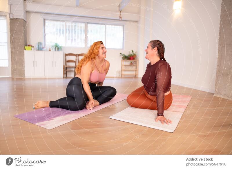 Fröhliche kurvige Frauen sitzen auf der Matte und lachen im Yoga-Zentrum heiter Lachen Talkrunde Lächeln diskutieren Sportbekleidung Pause Zusammensein üben