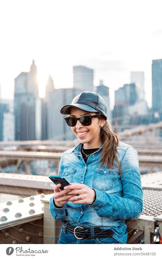 Frau mit Smartphone vor Manhattan-Kulisse Sonnenbrille Verschlussdeckel Lächeln Skyline Stadtbild New York State im Freien urban Brücke Mode Freizeit lässig
