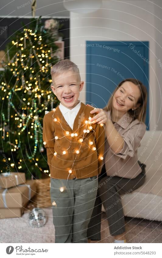 Fröhliches Kind mit seiner Mutter beim Spielen mit Weihnachtsbeleuchtung am Weihnachtsbaum zu Hause Junge lachen Weihnachten Lichter Freude Frau Feiertag