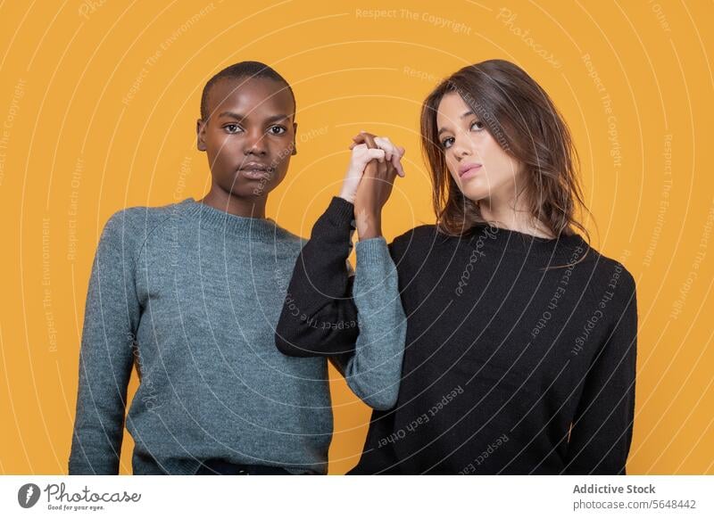 Verschiedene Frauen in Freizeitkleidung, die sich vor gelbem Hintergrund an den Händen halten selbstbewusst Freund Händchenhalten rassenübergreifend jung trendy