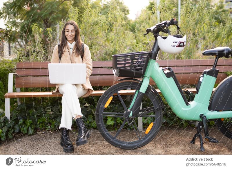 Junge Frau, die mit ihrem Elektrofahrrad im Park am Laptop arbeitet Bank sonnig im Freien arbeiten Tippen sitzend Freizeitkleidung Technik & Technologie