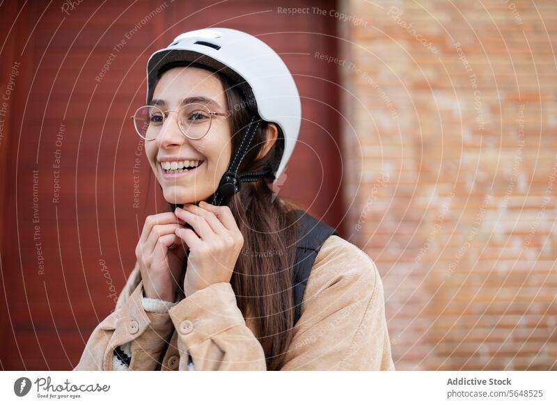 Lächelnde Frau befestigt Fahrradhelm im Freien Sicherheit Vorbereitung Gurt Sicherung Backsteinwand Mitfahrgelegenheit heiter jung Radfahrer Radfahren