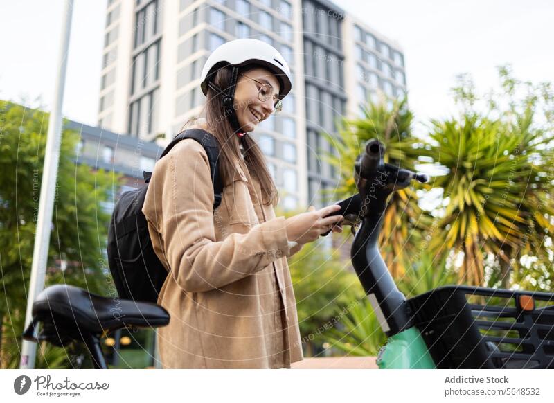 Junge Frau mit Helm und Smartphone auf einem E-Scooter in der Stadt Elektroroller urban Schutzhelm Freizeitkleidung Lächeln Stehen E-Roller Großstadt jung