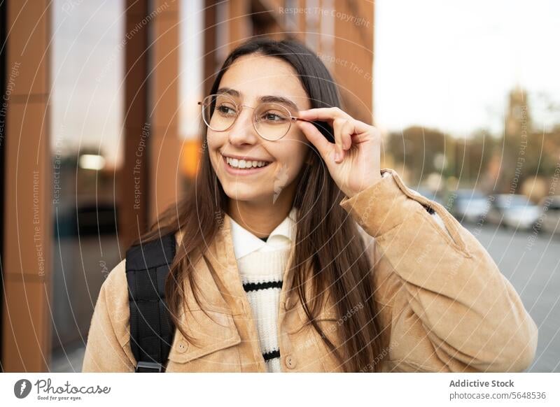 Lächelnde junge Frau, die ihre Brille auf einer Straße in der Stadt zurechtrückt Einstellung Großstadt Bürgersteig Stil Selbstvertrauen heiter urban lässig