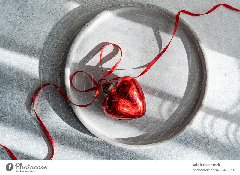 Draufsicht auf ein rotes Herz-Ornament, das auf einem Teller auf einer grauen Fläche liegt Hand rote Schleife Betonplatte Textur dynamisch Zuneigung