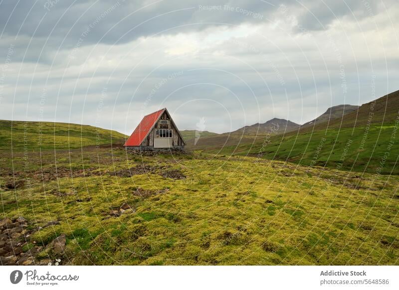 Von unten steht ein einsames Häuschen mit rotem Dach inmitten einer weiten isländischen Landschaft mit grünen Hügeln unter einem wolkenverhangenen Himmel Island