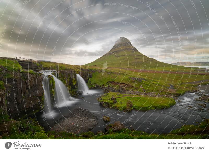 Üppige grüne Landschaft mit einem markanten Berggipfel, der sich über zwei Wasserfälle unter einem dramatischen Himmel in Island erhebt Berge u. Gebirge