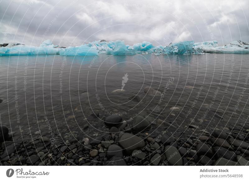 Eisberge schwimmen in einer Gletscherlagune unter einem bewölkten Himmel mit glatten Kieselsteinen im Vordergrund an der Küste in Island Lagune glazial wolkig