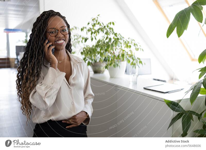 Nachdenkliche schwarze Geschäftsfrau mit Smartphone im Büro benutzend Lächeln Glück nachdenklich Hand auf Magen Afro-Look jung Business Arbeitsplatz Manager