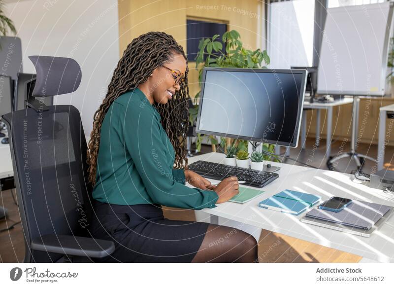 Seitenansicht einer jungen, glücklichen afroamerikanischen Geschäftsfrau, die an einem Computertisch im Büro auf einem Notizblock schreibt schreiben