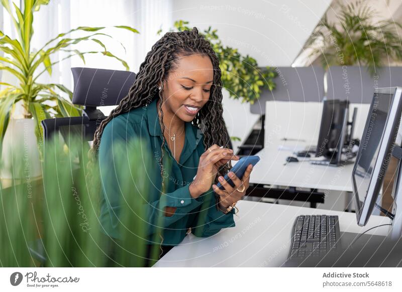 Glückliche schwarze Geschäftsfrau mit Smartphone am Schreibtisch im Büro benutzend Arbeit Business beschäftigt berührend Beruf Desktop pc Gerät Lächeln