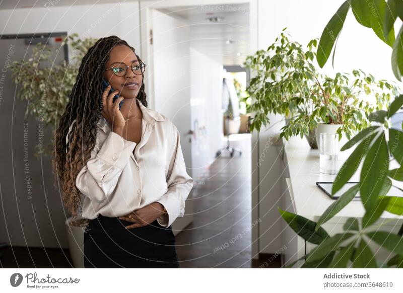 Nachdenkliche schwarze Geschäftsfrau mit Smartphone im Büro benutzend Lächeln Glück nachdenklich Hand auf Magen Afro-Look jung Business Arbeitsplatz Manager
