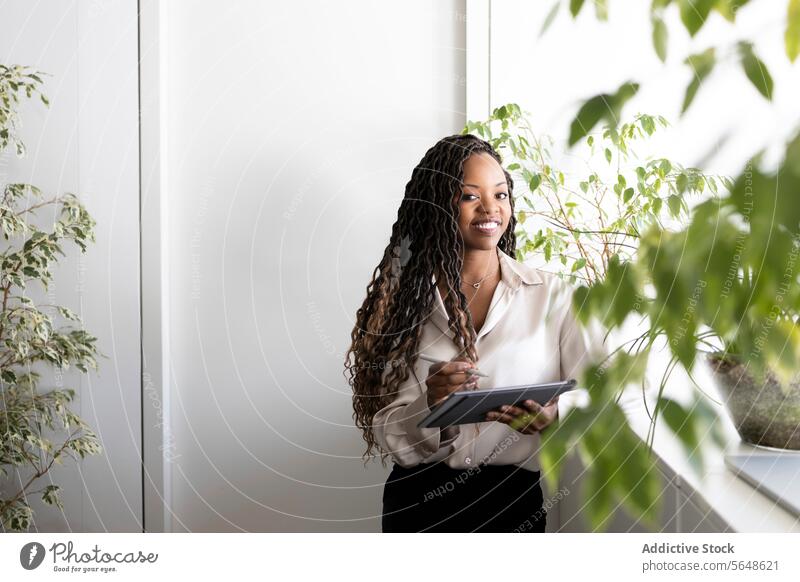 Nachdenkliche junge afroamerikanische Geschäftsfrau mit Faux-Locs-Frisur, die ein digitales Tablet und einen Stift hält, während sie in die Kamera schaut, während sie im Büro angibt