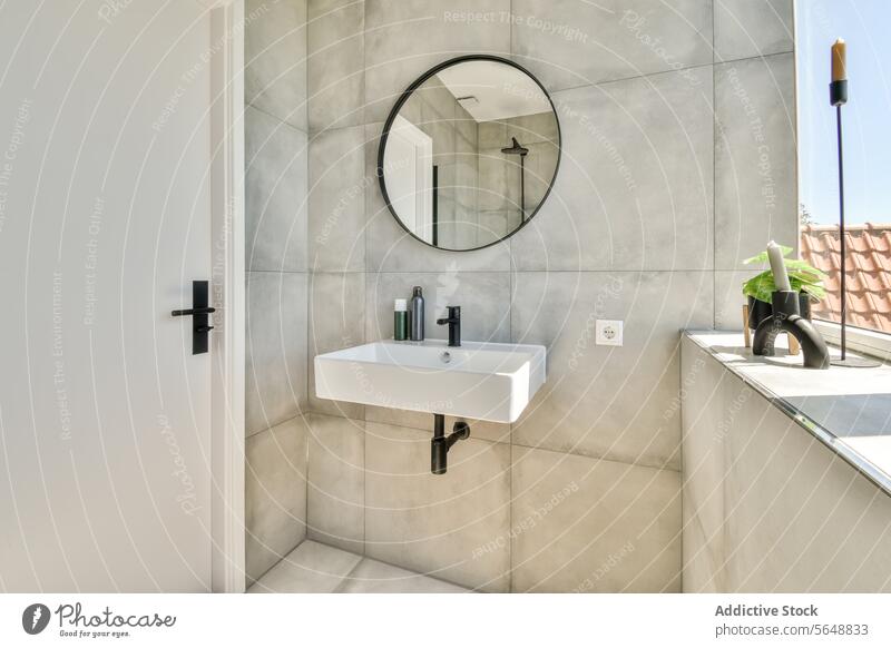 Interieur eines minimalistischen Badezimmers Spiegel Waschbecken Fenster Tür kreisrund montiert Wand weiß zugeklappt modern Sonnenlicht Kerze Textfreiraum