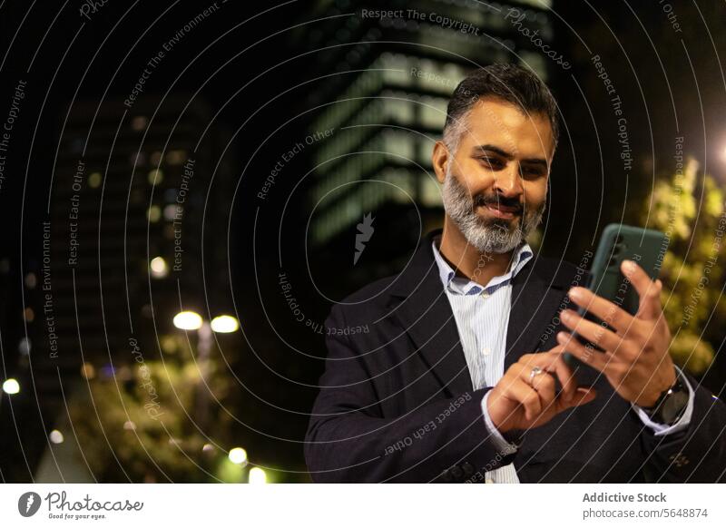 Glücklicher indischer Geschäftsmann im eleganten Anzug benutzt sein Smartphone auf einer Straße in der Stadt benutzend Funktelefon Lächeln Vollbart Stehen