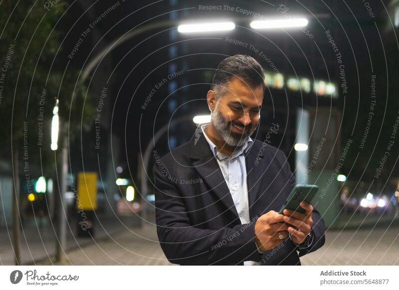 Glücklicher indischer Geschäftsmann im eleganten Anzug benutzt sein Smartphone auf einer Straße in der Stadt benutzend Funktelefon Lächeln Vollbart Stehen