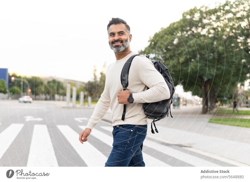 Glücklicher indischer Mann in Freizeitkleidung mit Rucksack schaut beim Überqueren der Straße weg Geschäftsmann Vollbart Großstadt Pullover Spaziergang klug