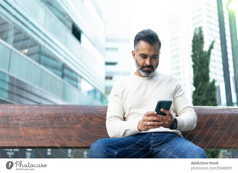 Konzentrierter indischer Geschäftsmann, der auf einer Straßenbank sitzend mit seinem Handy Nachrichten austauscht fokussiert Funktelefon Smartphone Jeanshose