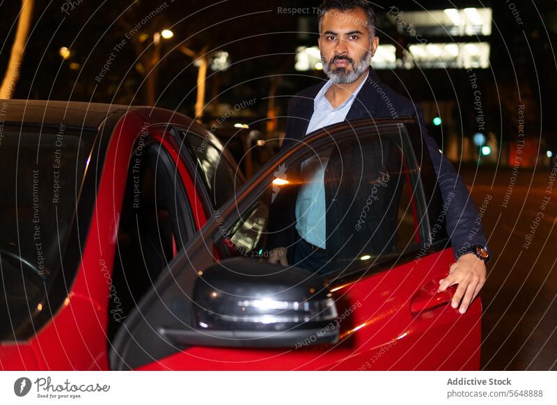 Seriöser ethnischer Fahrer steigt nachts auf der Straße in ein rotes Auto Geschäftsmann Automobil Laufwerk PKW Autoreise Mitfahrgelegenheit Unternehmer