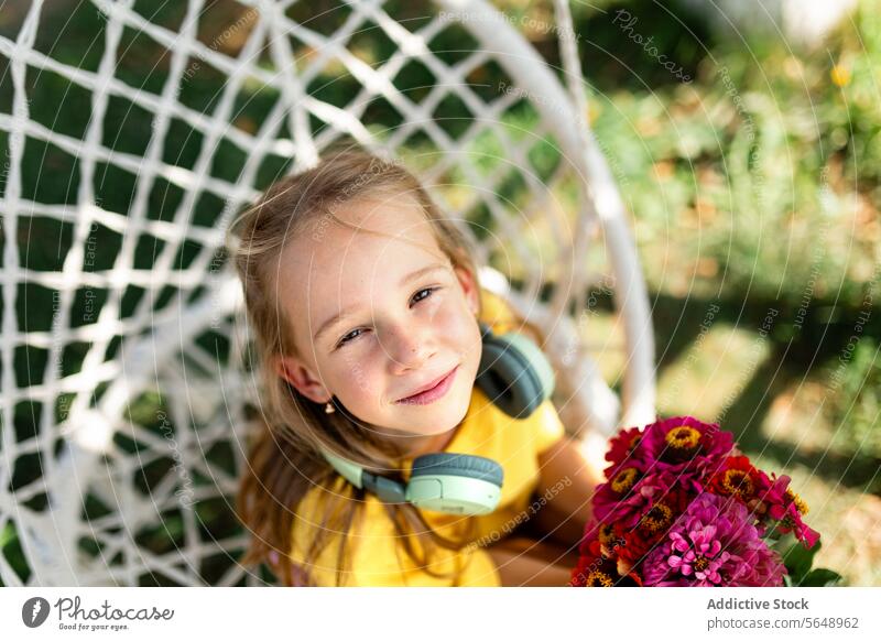 Junges Mädchen lächelnd mit Blumen in einem Hängesessel Kind Lächeln Glück Blumenstrauß Kopfhörer Stuhl Freizeit im Freien Garten Erholung freudig Jugend Natur